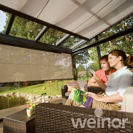 Paar sitzt entspannt im Wintergarten mit Sonnenschutz-Markise und blickt in den Garten.
